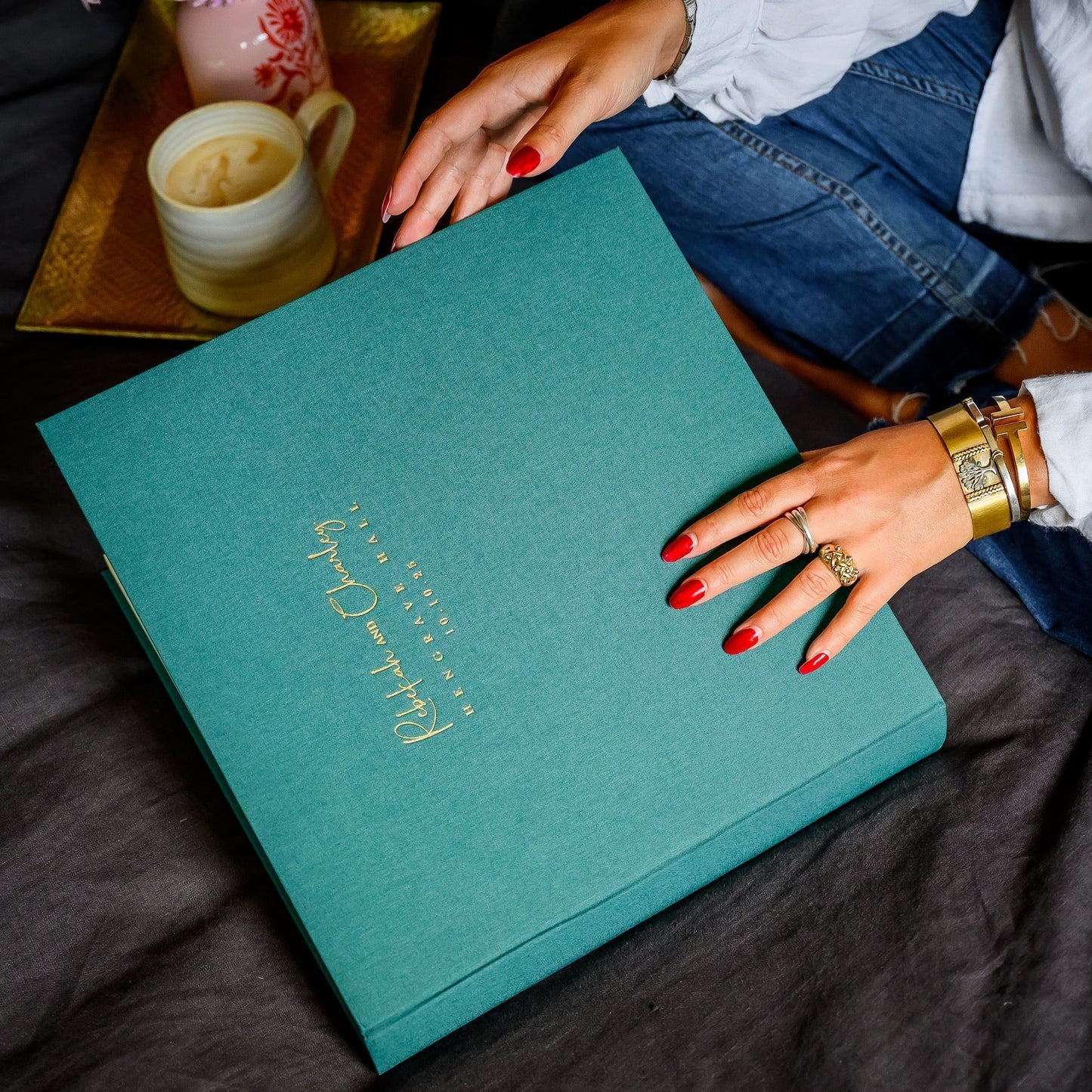 A woman is holding a green linen wedding photo album, poised to open it. The wedding album has been personalised on the front with gold writing. There is a cup of coffee in the backgound.