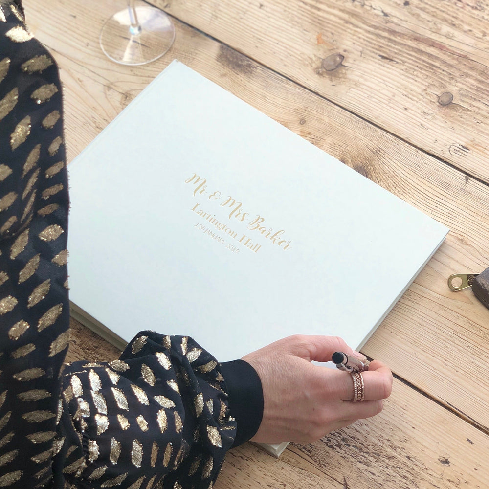 There is a landscape wedding guest book placed on a wooden table. A woman is holding a fountain pen about to leave a message in the guest book. 