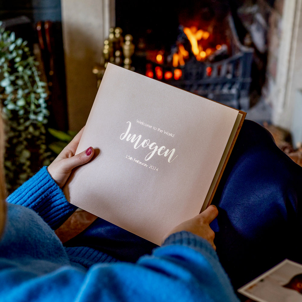 There is a woman in a blue jumper holding her baby's first album. It is a light pink album with a personalisation on the front. 