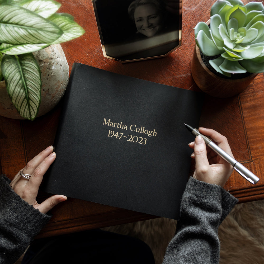 
                      
                        On a wooden table lies a black condolence book which has been printed with a woman's details in gold letters. There are some plants on the table and a photo of the deceased.
                      
                    