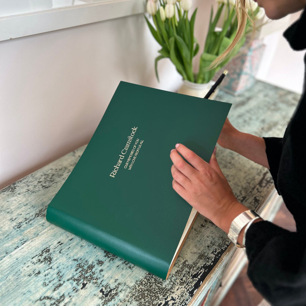 
                      
                        A large condolence book is on a sideboard and a woman is holding a pen writing a message inside it. There is a plant on the table too.
                      
                    