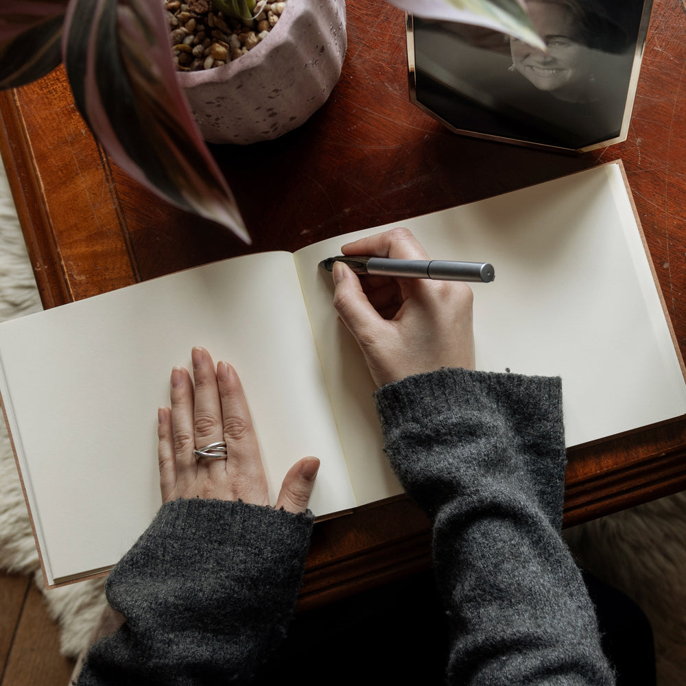 
                      
                        A large condolence book lies open on a wooden table and a woman in a grey jumper is poised with a pen and about to write a message inside it.
                      
                    