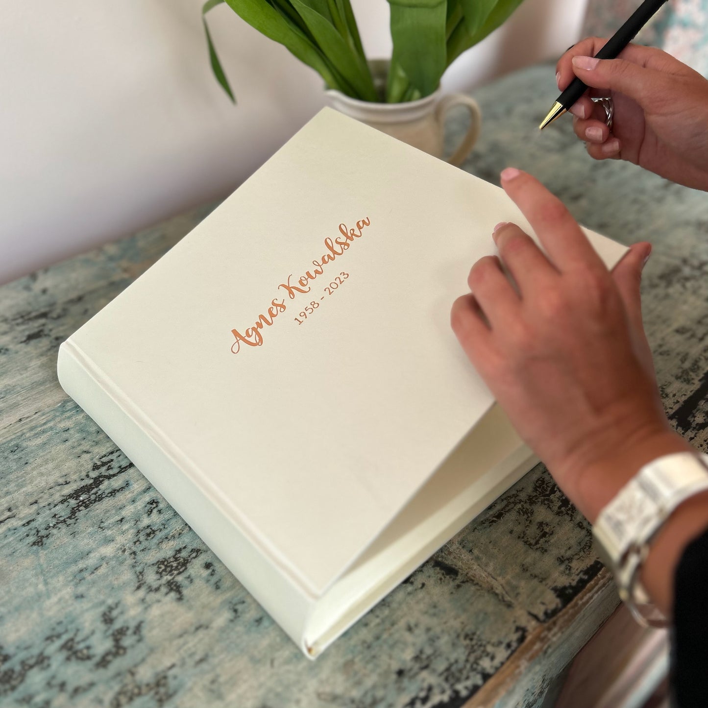 A large BeGolden condolence book is on a blue table. A woman is holding a pen ready to write a message inside.