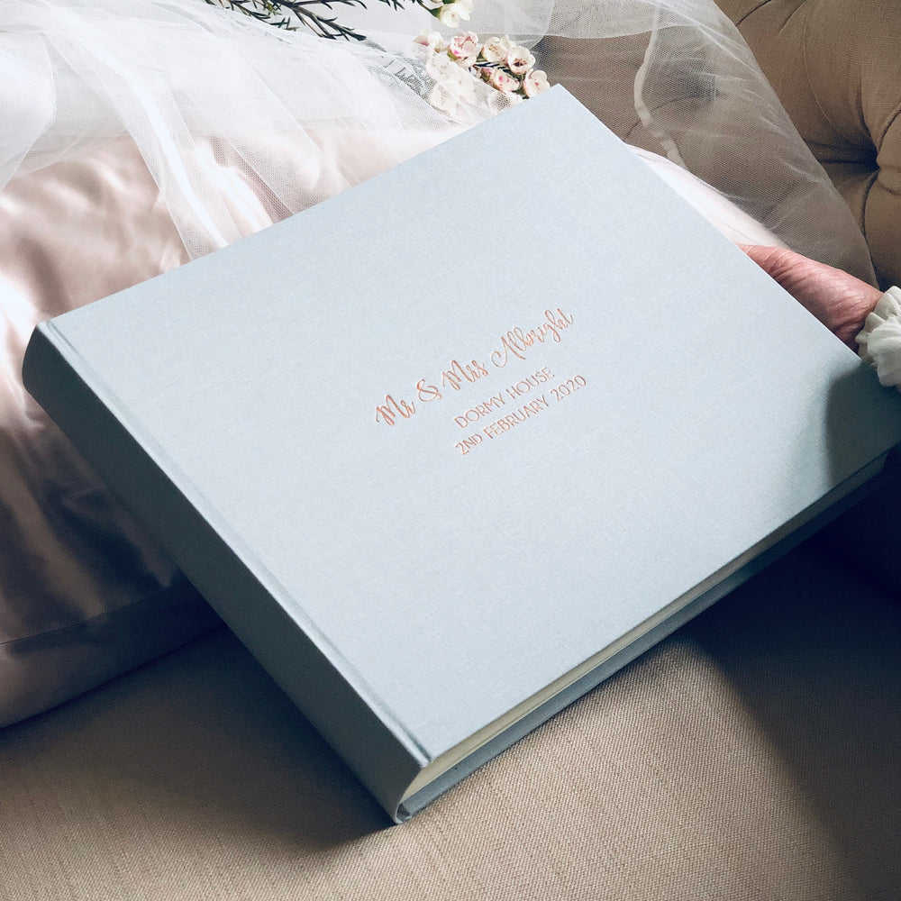 
                      
                        There is a large blue landscape linen wedding album in a woman's hand as she sits on a sofa. There are wedding details personalised on the front. 
                      
                    