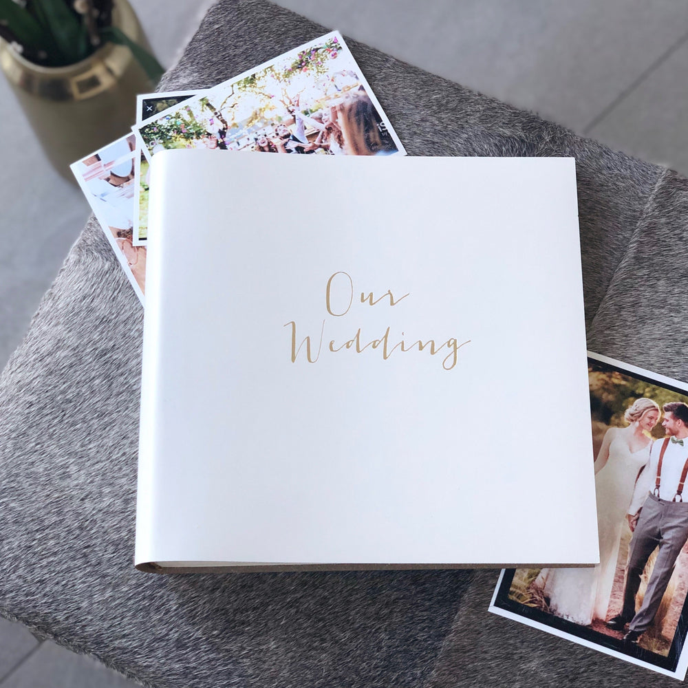 There is an ivory our wedding recycled leather album placed on a small table. There are photos surrounding the album ready to be placed inside.  