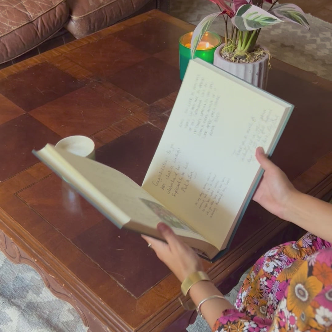 A woman is opening and then flicking through a guest book. She is reading the messages that her friends and family left in the book. There is a cup of coffee and a lit candle on the table. 