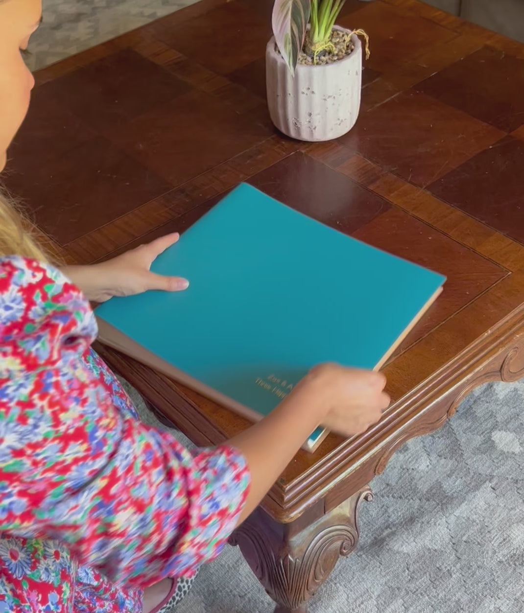 There is a woman who picks up an extra large album from a wooden table. She looks at the personalisation and then places it back on the table.  