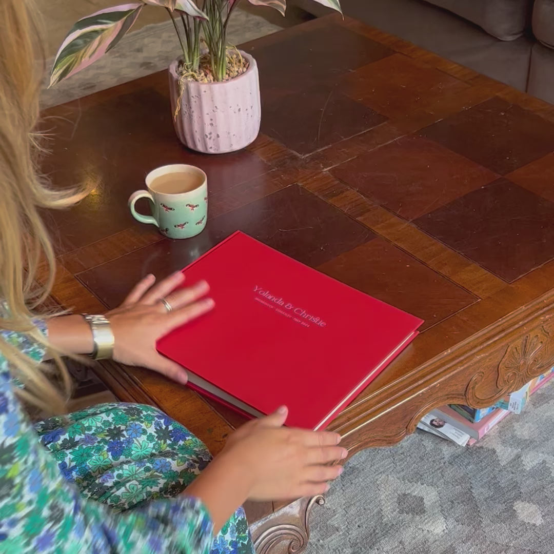 A girl picks up a large red album from her wooden coffee table, she looks at the front, which has been personalised with pink hot foil. She then turns the album and looks at the bind.