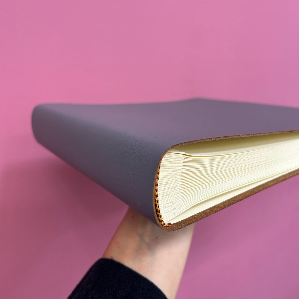 
                      
                        A grey condolence book is being held up against a pink background and you can see the pages inside.
                      
                    