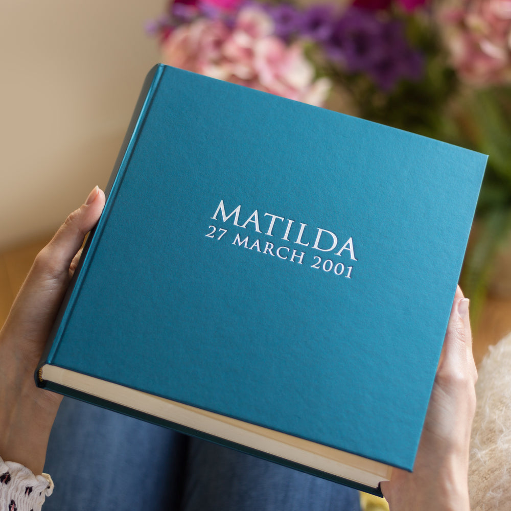 A close up image of a blue photo album which has been personalised with someone's name and date of birth. A large bunch of flowers is in the background.