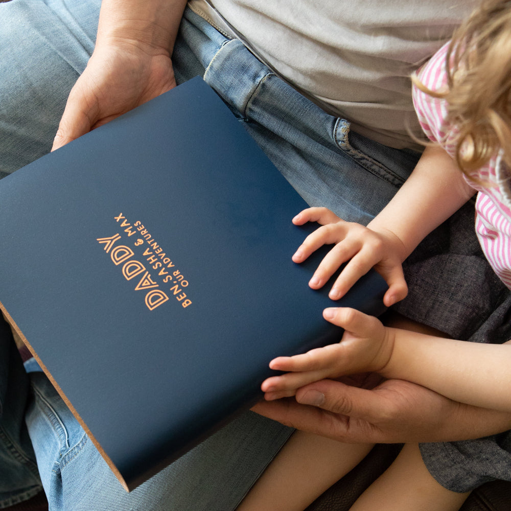 A daddy and his daughter are about to look through his personalised album. The album is in a dark blue shade and is a size medium. You can see both of their hands on the album. 