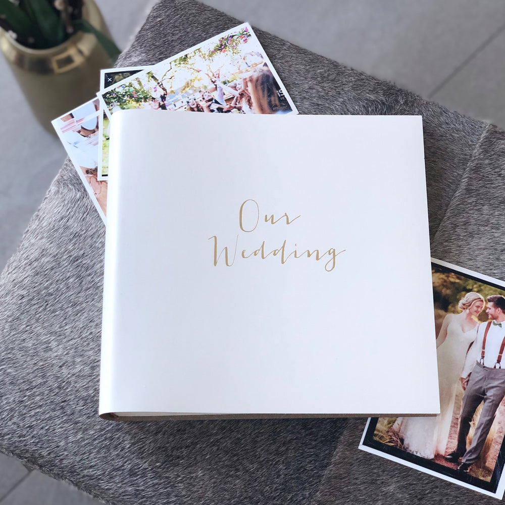 
                      
                        a square wedding album lies on a grey stool surrounded by wedding photos. The wedding album has been printed with Our Wedding on the front in gold foil
                      
                    