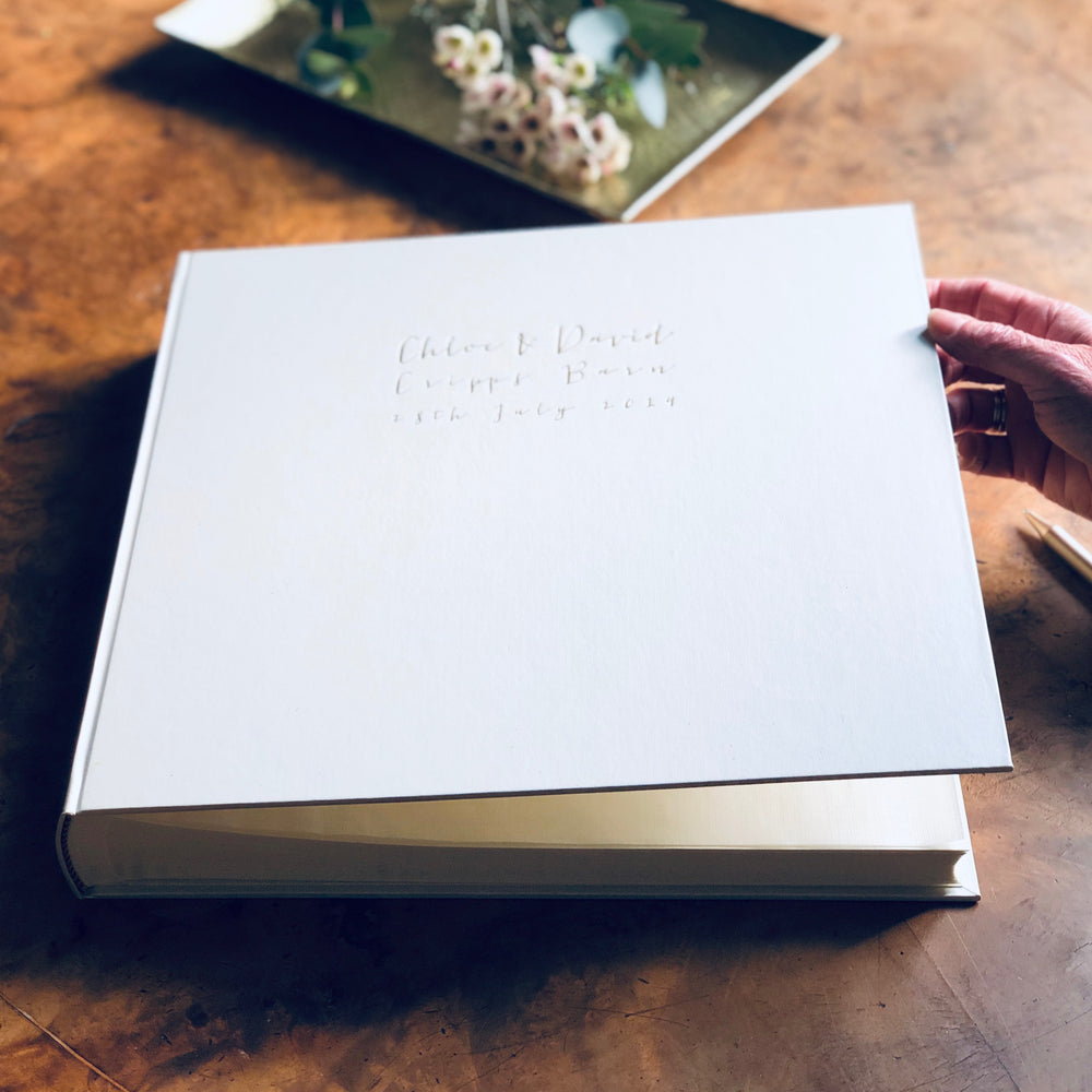 
                      
                        on a wooden table is a large ivory wedding album that has been personalised with wedding detals 
                      
                    