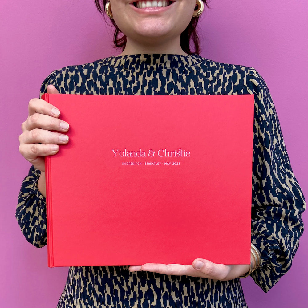 A woman in an animal print dress is standing in front of a pink wall. She is holding a red wedding guest book that has been personalised in a pink foil.