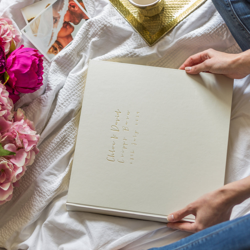 
                      
                        a large wedding photo album is on a white bed sheet and someone is holding it in their hands. There are some wedding photos also in the frame
                      
                    