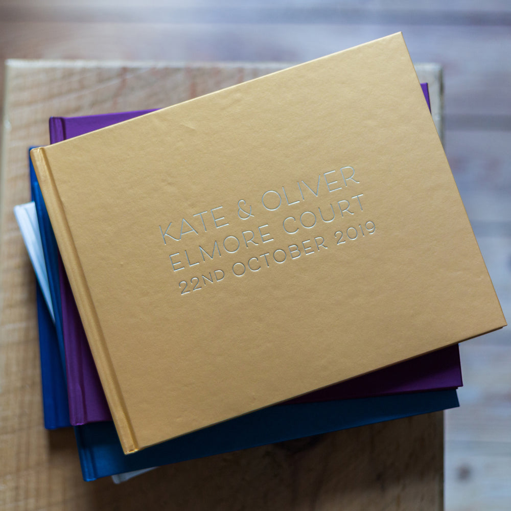  a pile of guest books are on a wooden table. the top one is a golden yellow an has wedding details printed on the front of it in a crisp font