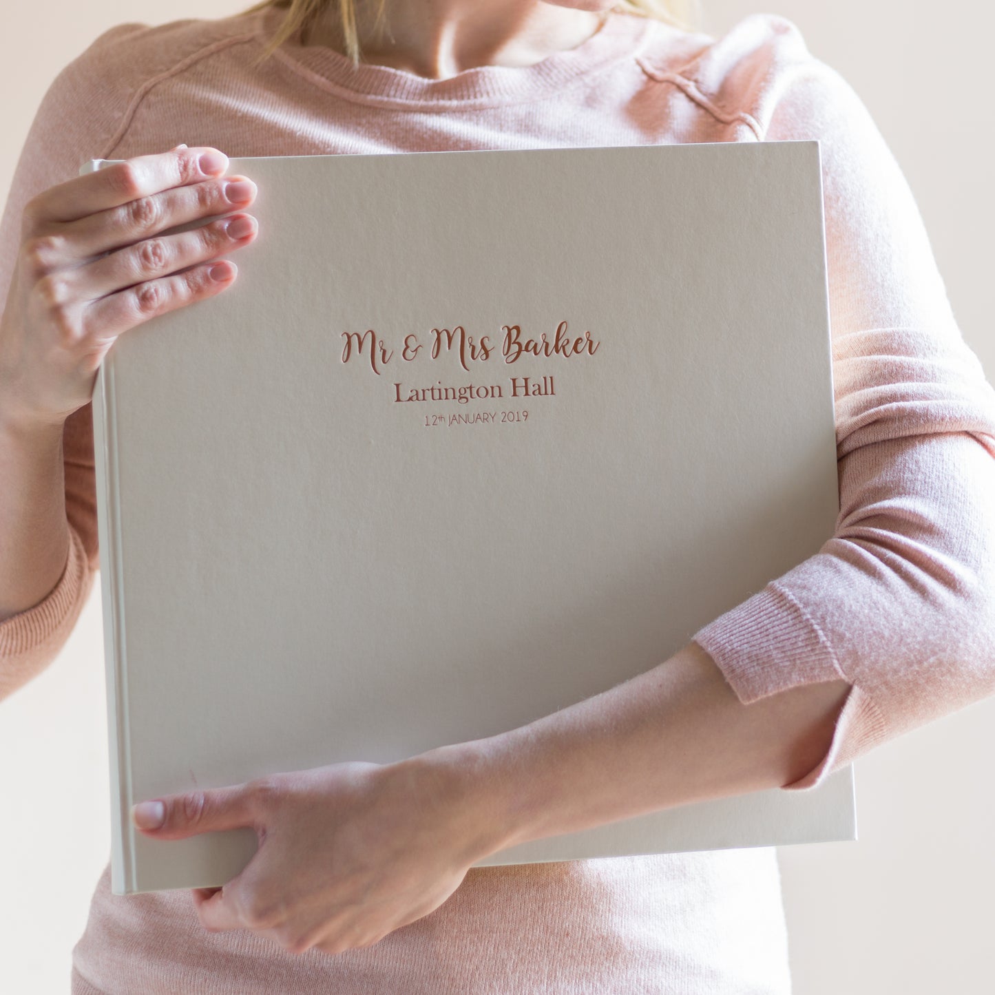 a girl in a pink jumper is clutching a large ivory wedding photo album. The photo album has wedding details hot foil printed onto the front.