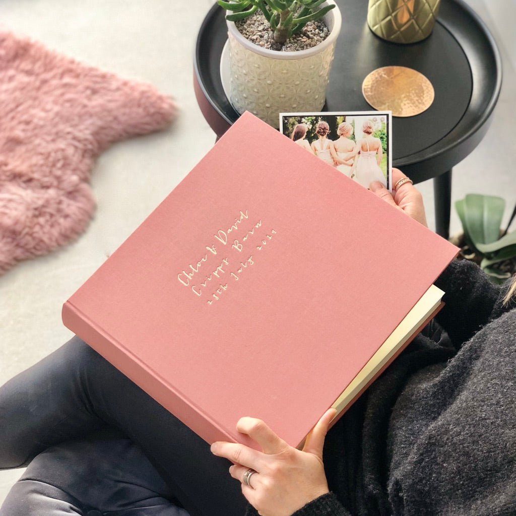 A woman is holding a large linen wedding album on her lap. A photograph is in her other hand and she is ready to place it inside the album. There is a small table also in shot, with a houseplant on it.