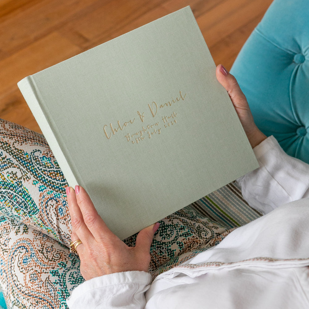 A woman is holding a large linen wedding guest book as she sits on a blue sofa. It has been personalised on the front with gold hot foil. 