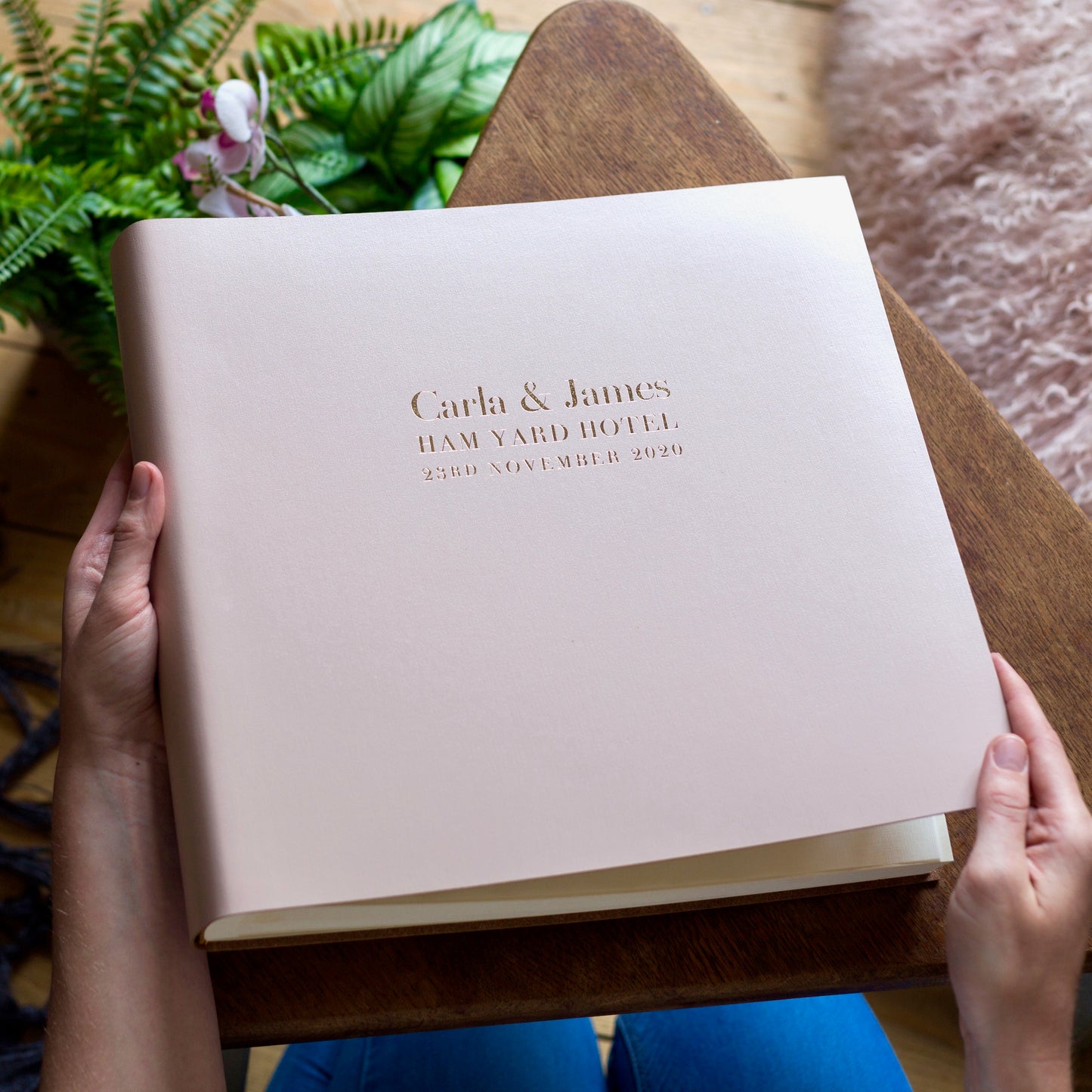 There is a large pale pink wedding album resting on a wooden table in a woman's hands. On the front of the album are her wedding details, printed with gold hot foil. 