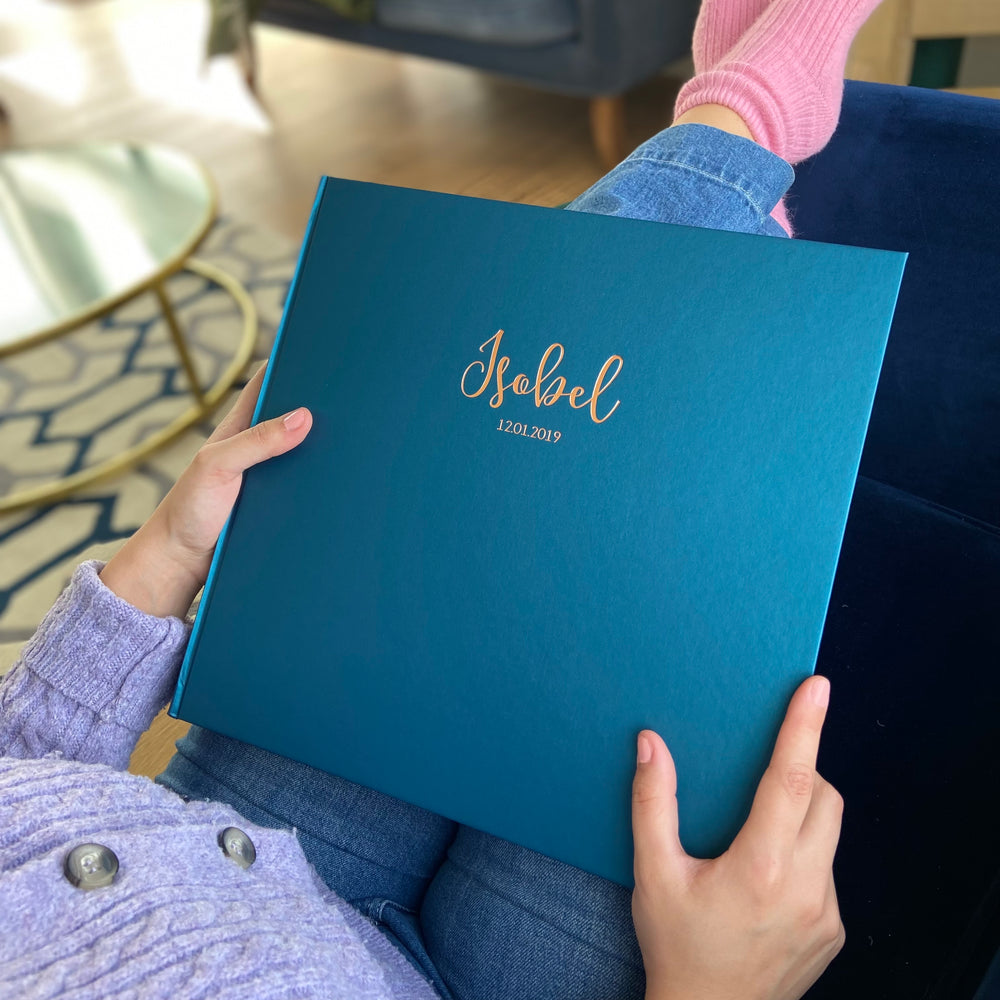 There is a woman sitting in a relaxed position on her sofa, she is holding her  daughters photo album which has been personalised with gold hot foil.