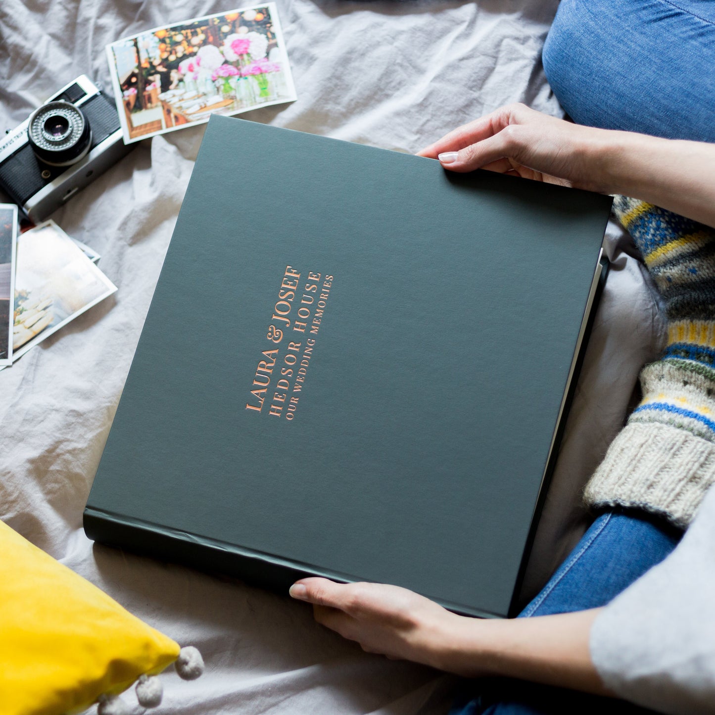 Someone is sitting on a bed holding a large black wedding photo album. The  album has been printed on the front with a bride and groom's wedding details in a copper foil. There is also a camera and a set of photographs on the bed.