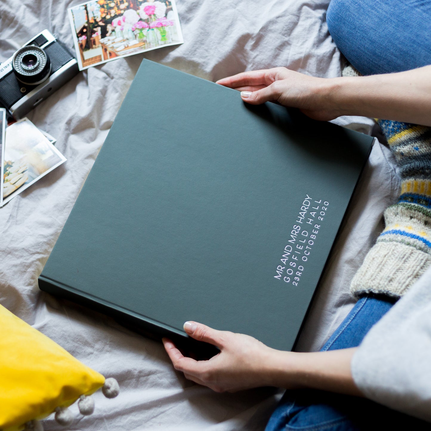 A large wedding album is being held in a woman's hands whilst she is sitting on her bed. The album has been printed along the bottom in a simple, sharp contemporary font.