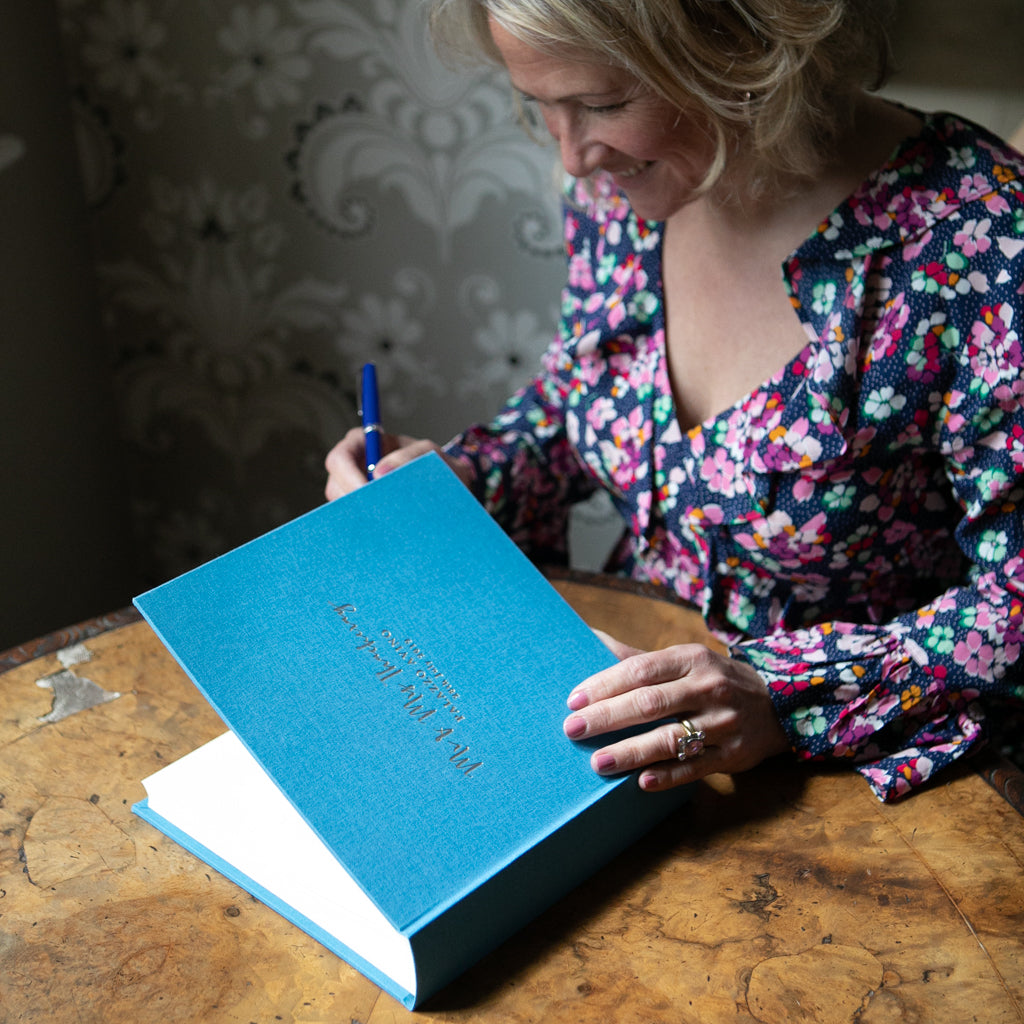 There is a woman in a floral dress who is about to write a message in a linen wedding book. It is in the colour blue and has been personalised on the front with gold hot foil.