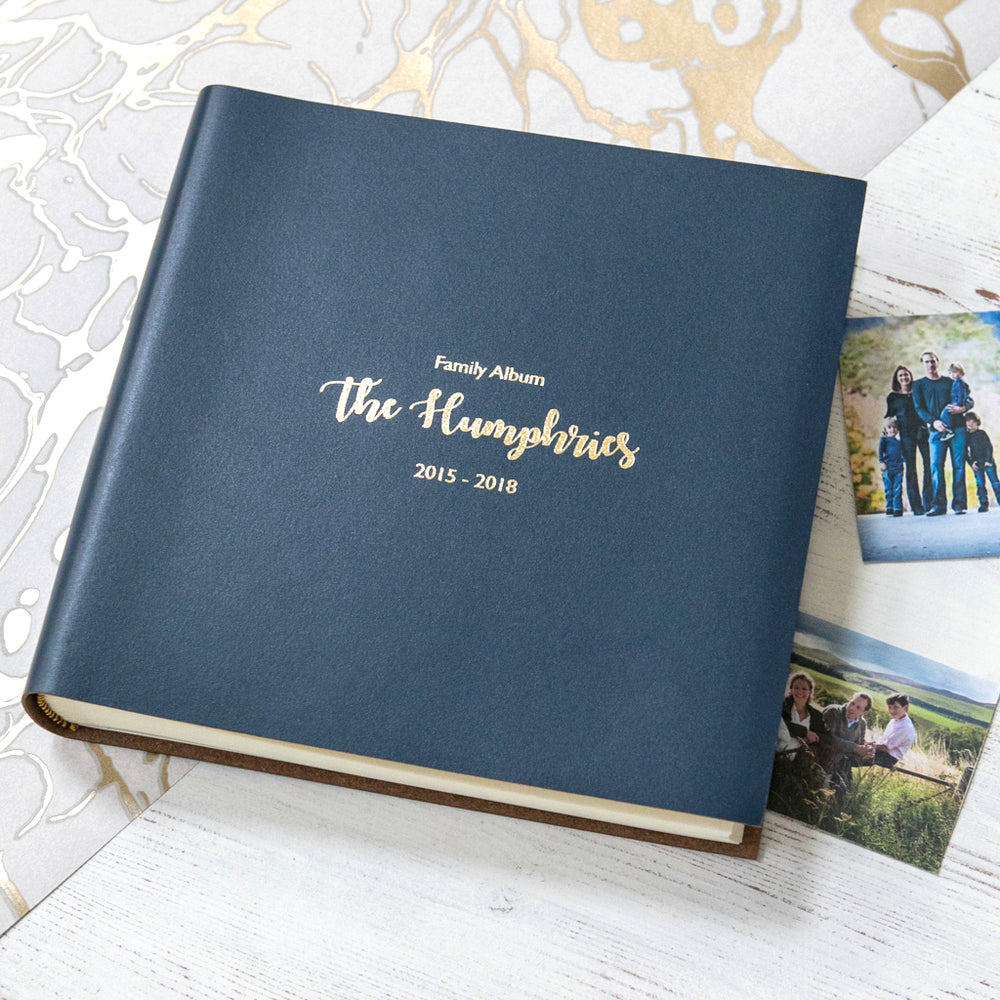 On a table lies a navy blue leather album which has been personalised with gold writing. This is a family album and you can see the photos which will be placed inside on the table too. 