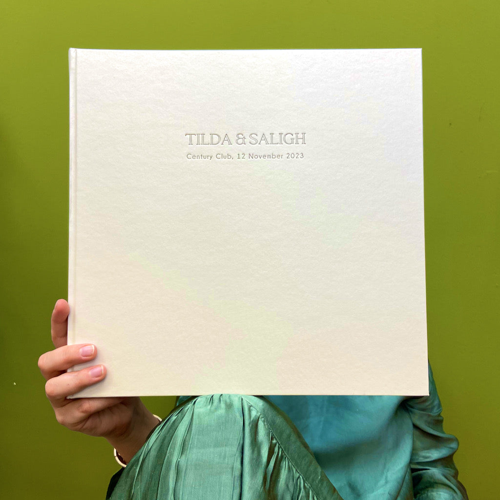 There is a woman holding up a large white jewel album. It has been personalised with someone's first names, a location and has also been dated. 