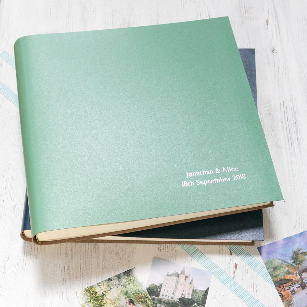 
                      
                        two large leather photo albums are on a table. The one on the top has been personalised with two lines of writing 
                      
                    