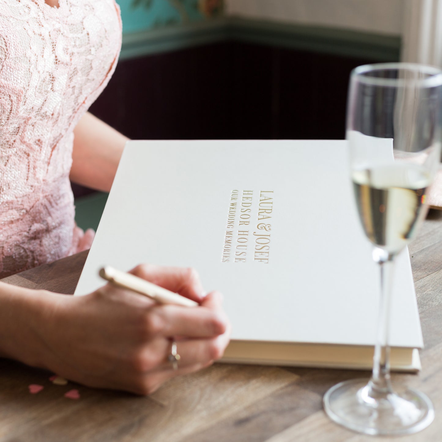 A wedding guest book is on a table and a guest is poised to write a message in it. She has a pen in her hand and there is a glass of Prosecco.  